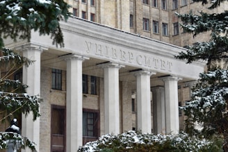 The entrance of a large, historic university building adorned with tall white columns and an inscription in Cyrillic above. The structure is made of light brick, and snow covers the trees and ground, capturing the essence of winter.