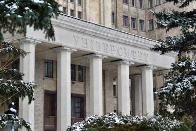 The entrance of a large, historic university building adorned with tall white columns and an inscription in Cyrillic above. The structure is made of light brick, and snow covers the trees and ground, capturing the essence of winter.