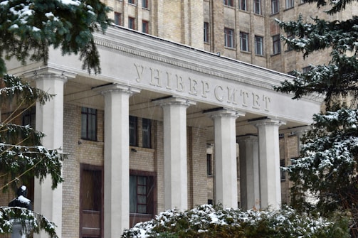 The entrance of a large, historic university building adorned with tall white columns and an inscription in Cyrillic above. The structure is made of light brick, and snow covers the trees and ground, capturing the essence of winter.
