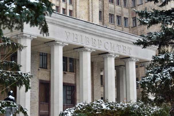 The entrance of a large, historic university building adorned with tall white columns and an inscription in Cyrillic above. The structure is made of light brick, and snow covers the trees and ground, capturing the essence of winter.