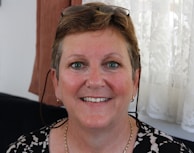 A middle-aged woman is smiling warmly. She has short, light brown hair and is wearing eyeglasses perched on her head. The background features a lace curtain and some brown fabric.