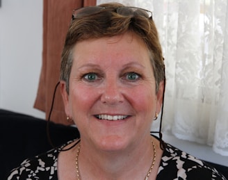 A middle-aged woman is smiling warmly. She has short, light brown hair and is wearing eyeglasses perched on her head. The background features a lace curtain and some brown fabric.