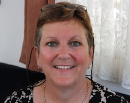 A middle-aged woman is smiling warmly. She has short, light brown hair and is wearing eyeglasses perched on her head. The background features a lace curtain and some brown fabric.