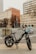 An electric bike is parked on the sidewalk in an urban setting, carrying a cooler and some colorful bags on the cargo racks. Buildings and leafless trees are visible in the background, along with traffic lights and a crosswalk.