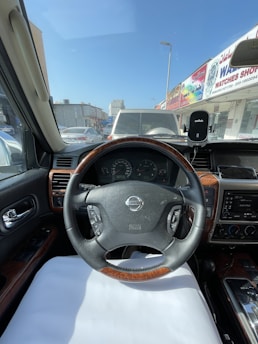 The interior of a car is shown with a focus on the steering wheel, which features a Nissan logo. There are controls on the steering wheel, and the dashboard is visible. Outside the windshield, several cars and a street with shops are seen. The driver's seat appears well-maintained with wood trim accents.