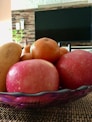 A bowl brimming with fresh fruits including apples, an orange, and what appears to be a mango, set on a woven mat. The setting appears to be indoors with a television in the background and a hint of houseplants at the side.