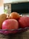 A bowl brimming with fresh fruits including apples, an orange, and what appears to be a mango, set on a woven mat. The setting appears to be indoors with a television in the background and a hint of houseplants at the side.