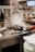 A kitchen scene with a person preparing food. Steam rises from a pot filled with mussels, and various kitchen utensils and ingredients surround the area. A hand is holding a pan, and plates are stacked nearby.