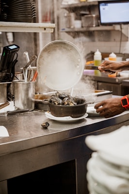A kitchen scene with a person preparing food. Steam rises from a pot filled with mussels, and various kitchen utensils and ingredients surround the area. A hand is holding a pan, and plates are stacked nearby.