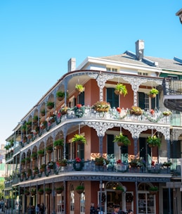 A historic building with ornate wrought-iron balconies adorned with hanging plants and decorations, featuring multiple stories and a corner position. The architecture displays a blend of brickwork and intricate metalwork, typical of New Orleans-style structures.