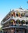 A historic building with ornate wrought-iron balconies adorned with hanging plants and decorations, featuring multiple stories and a corner position. The architecture displays a blend of brickwork and intricate metalwork, typical of New Orleans-style structures.