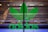 A large green Adidas logo is displayed on a glass surface. The background consists of a blurred, metallic fence or grille which gives an industrial look.