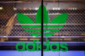 A large green Adidas logo is displayed on a glass surface. The background consists of a blurred, metallic fence or grille which gives an industrial look.