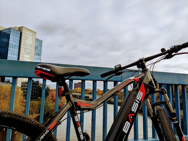 A bicycle with the brand name BASIS prominently displayed on the frame is propped against a blue railing of a bridge. In the background, there are modern multi-story buildings with reflective glass windows and a river or large body of water surrounded by trees with autumn foliage.