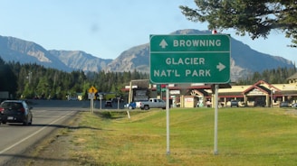 A road sign indicating directions to Browning and Glacier National Park stands next to a highway. Behind the sign, there are cars on the road and a strip of shops with red roofs. In the background, a range of mountains under a clear blue sky is visible.