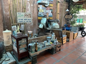 A vintage-style coffee shop setting features a collection of eclectic items. A retro scooter is mounted as part of the decor against a wall adorned with license plates and rustic signs. There's a mix of old-fashioned furniture, including a small wooden cabinet filled with various memorabilia and bottles on display. The scene also includes a table set up with coffee cups, bottles, and snacks. Plants and bamboo add a touch of greenery to the space, creating a nostalgic and cozy atmosphere.