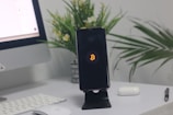 A smartphone with a Bitcoin logo on the screen is placed on a black stand. Nearby are a computer monitor, a white keyboard, a closed charging case for earbuds, and a pen. In the background, there are potted green plants with small yellow and white flowers.