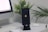 A smartphone with a Bitcoin logo on the screen is placed on a black stand. Nearby are a computer monitor, a white keyboard, a closed charging case for earbuds, and a pen. In the background, there are potted green plants with small yellow and white flowers.