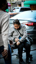An elderly person is sitting on a bench in an urban setting, wearing a grey jacket and black pants. They're holding something, perhaps adjusting or pouring it, with a focused expression on their face. Cars and people move in the background, suggesting that this is a busy street scene.