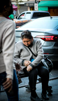 An elderly person is sitting on a bench in an urban setting, wearing a grey jacket and black pants. They're holding something, perhaps adjusting or pouring it, with a focused expression on their face. Cars and people move in the background, suggesting that this is a busy street scene.