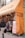 A storefront with a bright orange awning featuring text in Japanese. Bedding products, including pillows and quilts, are displayed outside on racks. A woman with long hair, wearing a black jacket, holds a dark-colored bag as she walks past the store.