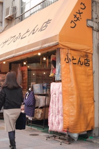 A storefront with a bright orange awning featuring text in Japanese. Bedding products, including pillows and quilts, are displayed outside on racks. A woman with long hair, wearing a black jacket, holds a dark-colored bag as she walks past the store.