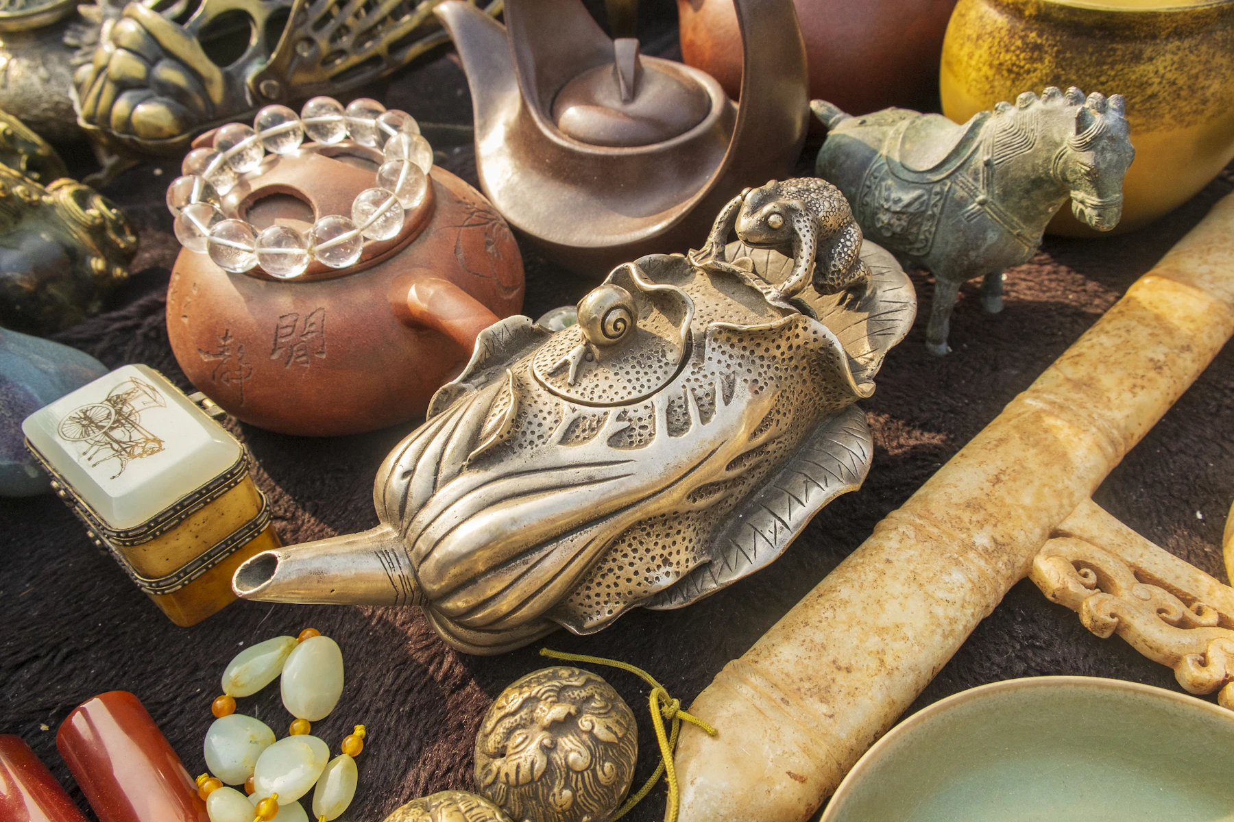 A flea market table crowded with a silver-colored teapot shaped like a cabbage leaf, a red clay teapot, beads and carvings