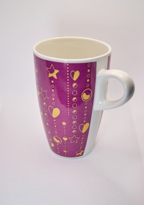 A ceramic mug with a tall, cylindrical shape and a handle on one side. The mug features a purple background adorned with yellow decorative elements, including stars, hearts, and circles arranged in vertical lines.