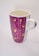 A ceramic mug with a tall, cylindrical shape and a handle on one side. The mug features a purple background adorned with yellow decorative elements, including stars, hearts, and circles arranged in vertical lines.