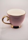 A glossy pink ceramic cup with a gold handle, rim, and base, set against a neutral background.