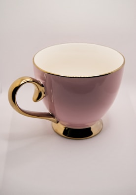 A glossy pink ceramic cup with a gold handle, rim, and base, set against a neutral background.