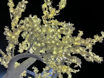 A close-up view of an artificial tree with numerous branches adorned with bright white leaves and twinkling lights against a dark sky. The lights are evenly distributed, illuminating the leaves and giving a glowing appearance.