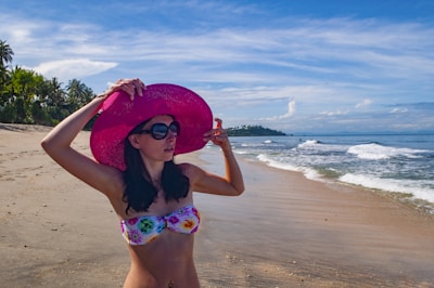 A woman stands on a sandy beach wearing a floral bikini and a large pink sun hat, adjusting her sunglasses. The beach is lined with green palm trees, and the ocean waves gently roll onto the shore under a clear blue sky with scattered clouds.