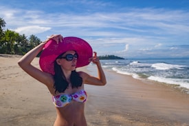 A woman stands on a sandy beach wearing a floral bikini and a large pink sun hat, adjusting her sunglasses. The beach is lined with green palm trees, and the ocean waves gently roll onto the shore under a clear blue sky with scattered clouds.