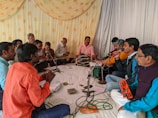 A group of men are sitting in a circle, engaging in a musical performance. They are holding various musical instruments such as microphones, a harmonium, and a small tambourine. The setting appears to be indoors with a decorative curtain featuring golden patterns in the background. Various sheets of paper are visible on the floor, and a white sheet covers the ground where the men are seated.