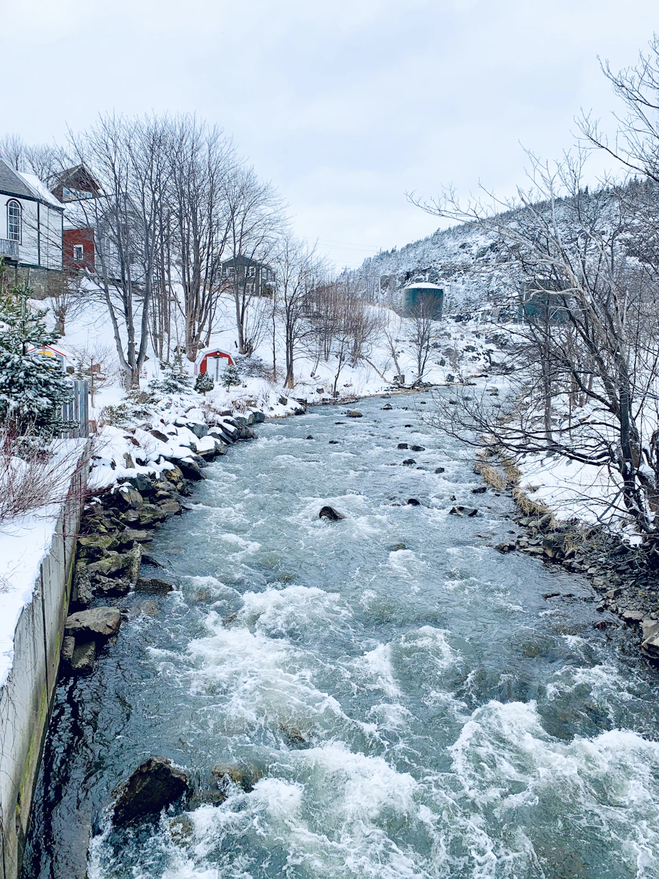 The image depicts a serene winter scene at a riverbank, with snow-covered trees and houses lining the banks. The river is flowing rapidly through the center of the frame, its white waters contrasting against the surrounding landscape. On either side of the river, there are two houses - one on each bank. In the foreground, there's a small red building that stands out against the snowy backdrop.