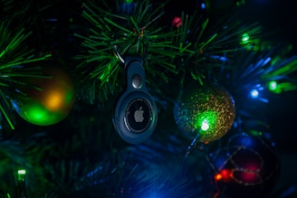 A Christmas tree adorned with colorful lights and ornaments, featuring a prominently displayed electronic tag with a logo in the center. The surrounding ornaments include a green and gold bauble and a glittering red bauble, all illuminated by the vibrant green and blue lights.