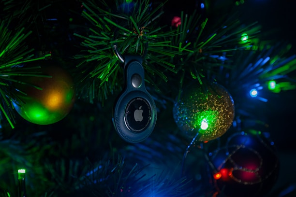 A Christmas tree adorned with colorful lights and ornaments, featuring a prominently displayed electronic tag with a logo in the center. The surrounding ornaments include a green and gold bauble and a glittering red bauble, all illuminated by the vibrant green and blue lights.