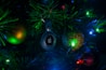 A Christmas tree adorned with colorful lights and ornaments, featuring a prominently displayed electronic tag with a logo in the center. The surrounding ornaments include a green and gold bauble and a glittering red bauble, all illuminated by the vibrant green and blue lights.