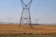 A rural landscape featuring multiple electricity pylons and power lines spanning across an open, dry field. The scene extends into the horizon with a clear blue sky overhead, and scattered trees can be seen in the distance.