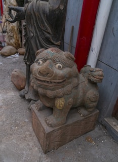 A stone or ceramic statue of a mythical creature, resembling a lion or dragon, is situated on a carved pedestal. The creature features exaggerated facial expressions with large eyes and a wide mouth. Beside it stands a larger robed figure, possibly another statue, with its hand partially visible pointing outward. The area appears to be an outdoor setting or workshop with other stone sculptures and materials visible in the background.