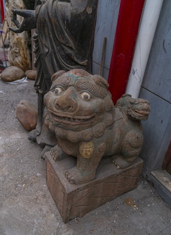 A stone or ceramic statue of a mythical creature, resembling a lion or dragon, is situated on a carved pedestal. The creature features exaggerated facial expressions with large eyes and a wide mouth. Beside it stands a larger robed figure, possibly another statue, with its hand partially visible pointing outward. The area appears to be an outdoor setting or workshop with other stone sculptures and materials visible in the background.