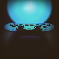 A video game controller is prominently featured against an illuminated background with a gradient of blue light. The controller appears to be of a modern design with dual joysticks and multiple buttons visible. The focus is on the upper section of the controller, creating a contrast between the lighted background and the shadowed device.