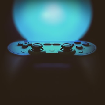 A video game controller is prominently featured against an illuminated background with a gradient of blue light. The controller appears to be of a modern design with dual joysticks and multiple buttons visible. The focus is on the upper section of the controller, creating a contrast between the lighted background and the shadowed device.