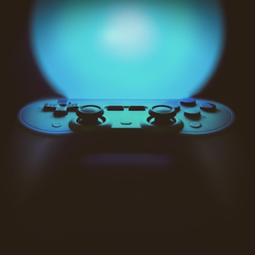 A video game controller is prominently featured against an illuminated background with a gradient of blue light. The controller appears to be of a modern design with dual joysticks and multiple buttons visible. The focus is on the upper section of the controller, creating a contrast between the lighted background and the shadowed device.