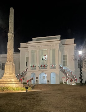 A historic white courthouse building, established in 1822, is beautifully decorated for the holiday season with red bows and lights along the railings and the building facade. An obelisk monument stands prominently to the left of the courthouse, illuminated by ground lighting. The scene is set at night, highlighting the festive decorations and architectural features.