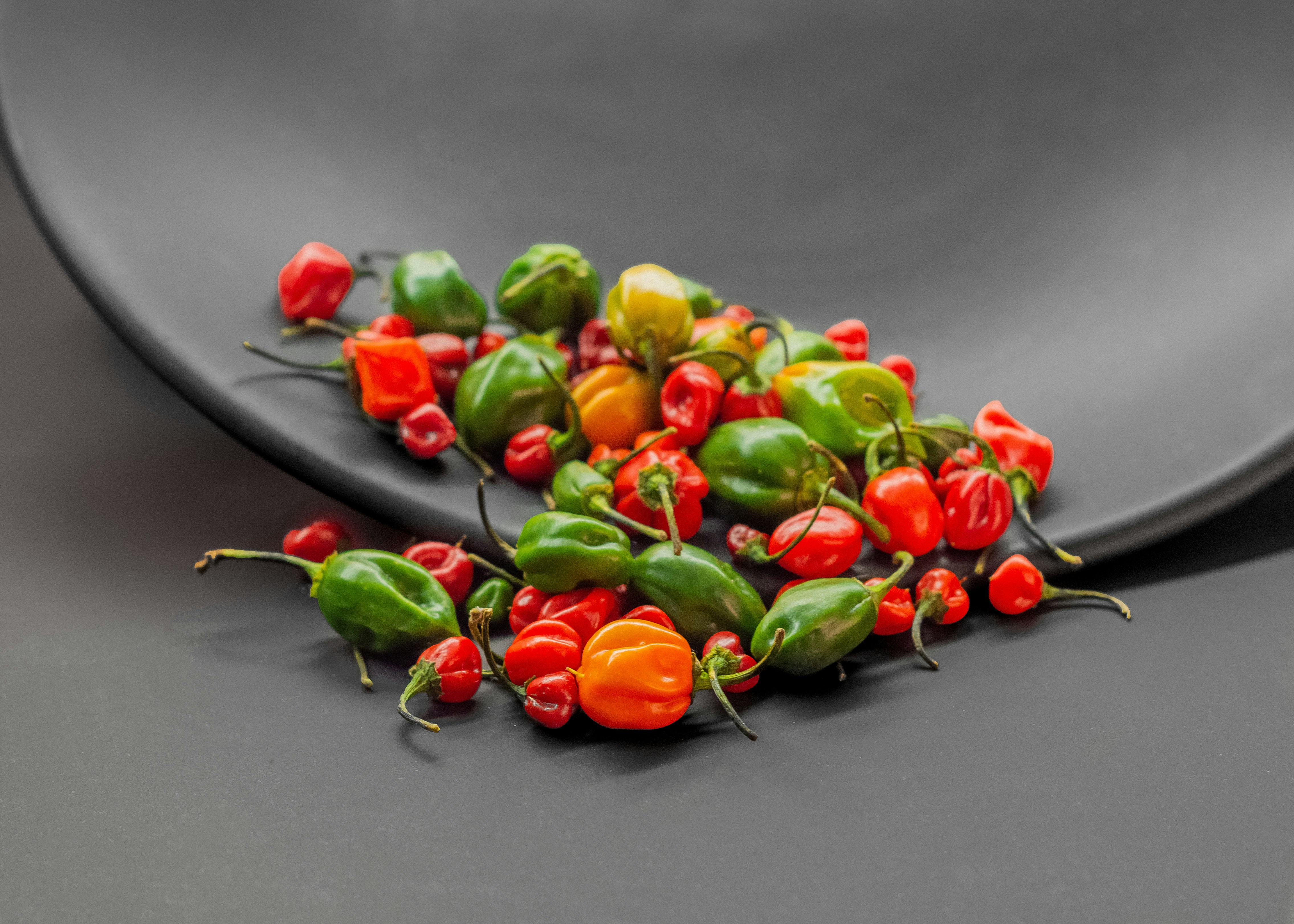 a pile of peppers sitting on top of a black plate, Habanero chili peppers falling on isolated white background e mexico, Naga jolokia (the hottest chili peppers in the world)