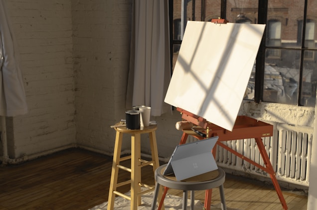 A bright studio room with brick walls and wooden floors features a large window. Sunlight streams in, casting shadows on a blank canvas mounted on an easel. Nearby, a wooden stool holds several paint cans, and a laptop is placed on a small round table.