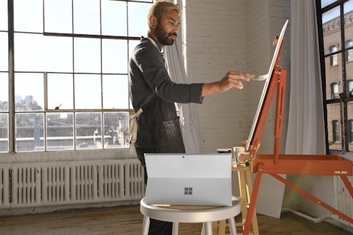 A person stands by an easel, painting in a well-lit studio with large windows. The interior is modern, featuring white brick walls and a wooden floor. Natural light streams through the windows, creating a warm and inviting atmosphere. A tablet is placed on a small round table nearby.
