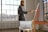 A person stands by an easel, painting in a well-lit studio with large windows. The interior is modern, featuring white brick walls and a wooden floor. Natural light streams through the windows, creating a warm and inviting atmosphere. A tablet is placed on a small round table nearby.